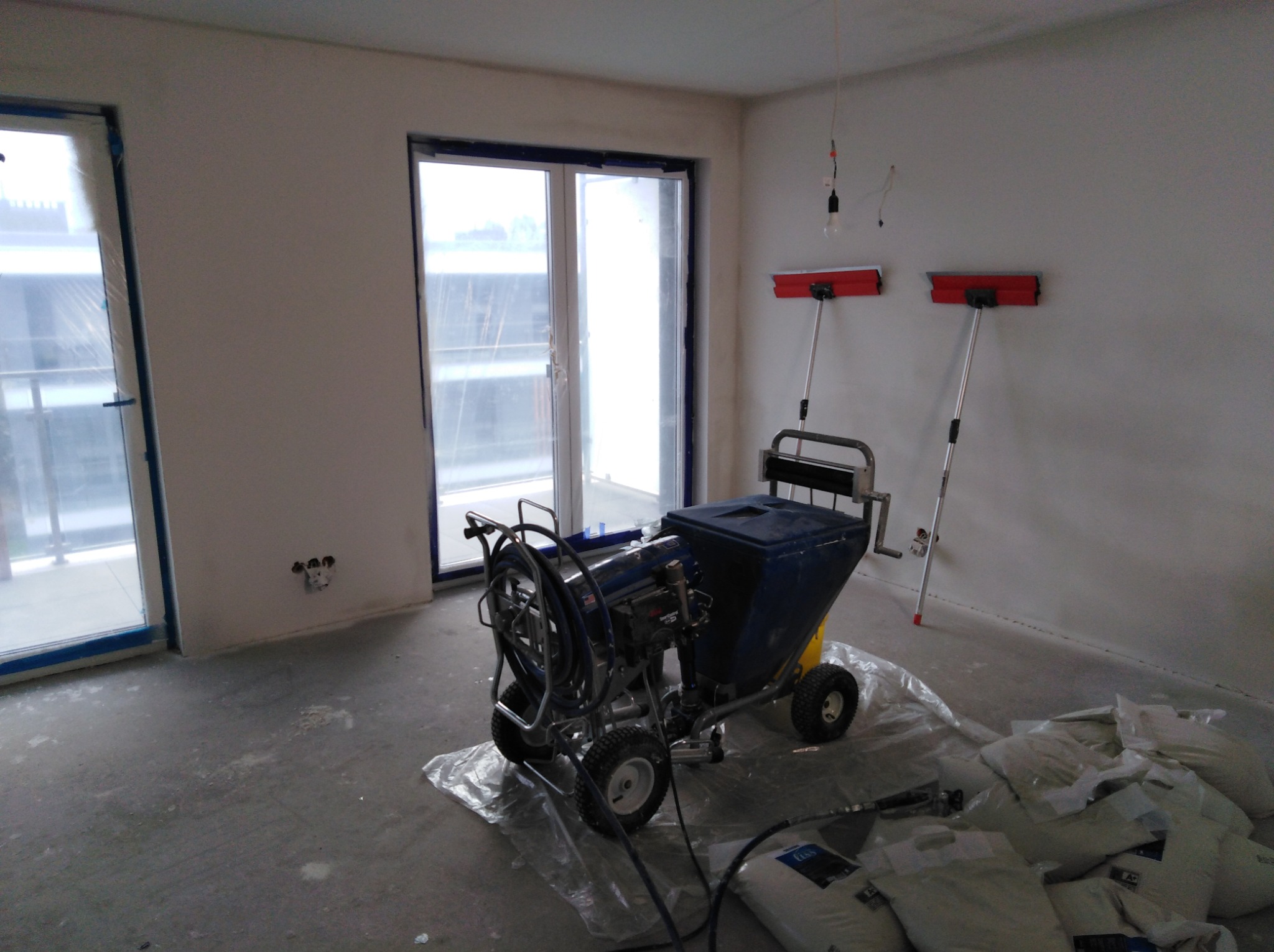 Przygotowanie do szpachlowania maszynowego: agregat szpachlarski na folii, worki z gładzią oraz długie szpachelki przy ścianie w surowym wnętrzu z zabezpieczonymi oknami.