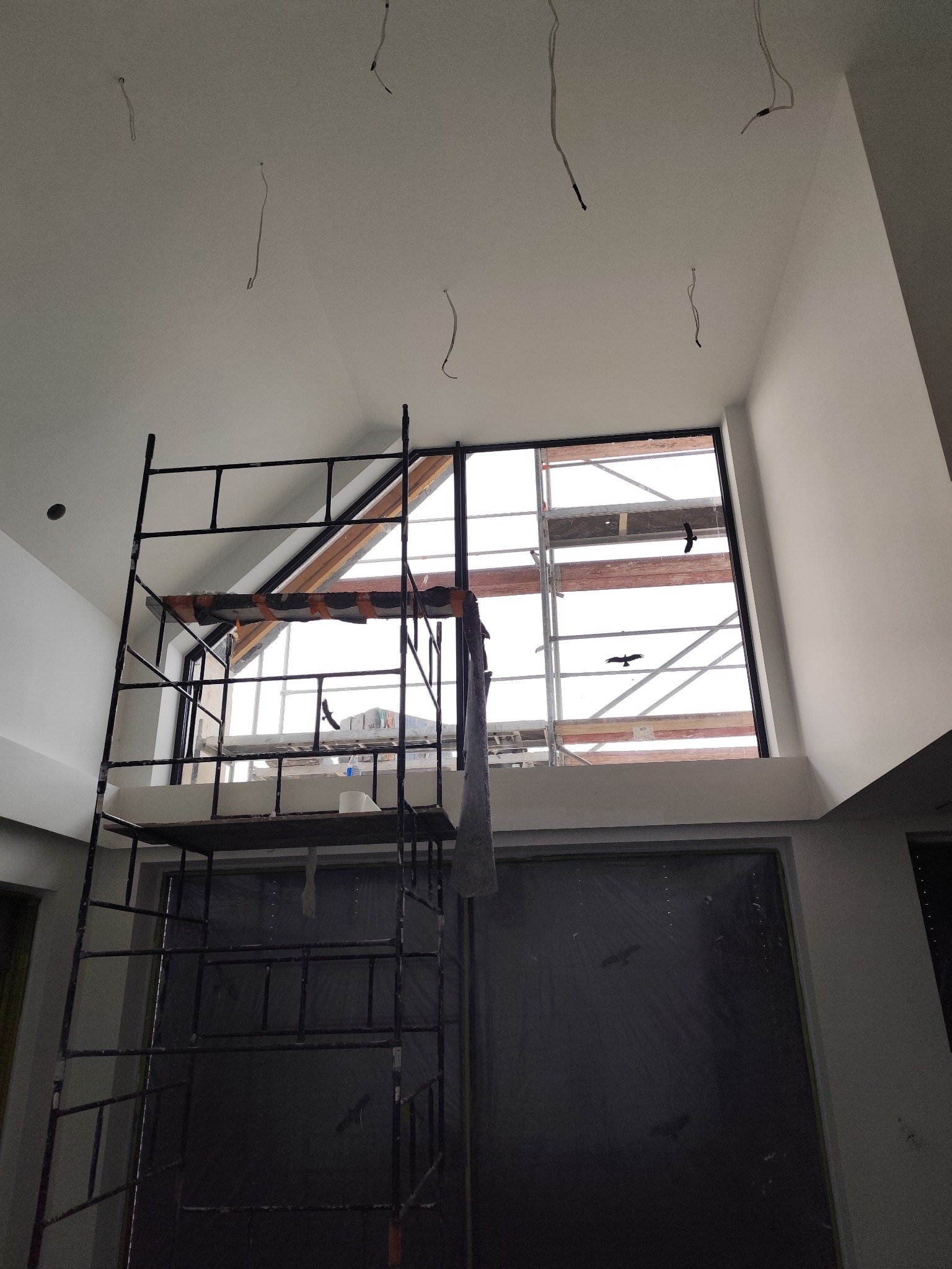 Wnętrze w trakcie prac wykończeniowych: rusztowanie, okno z widokiem na zewnątrz, wystające kable z sufitu. Ściany przygotowane do malowania.