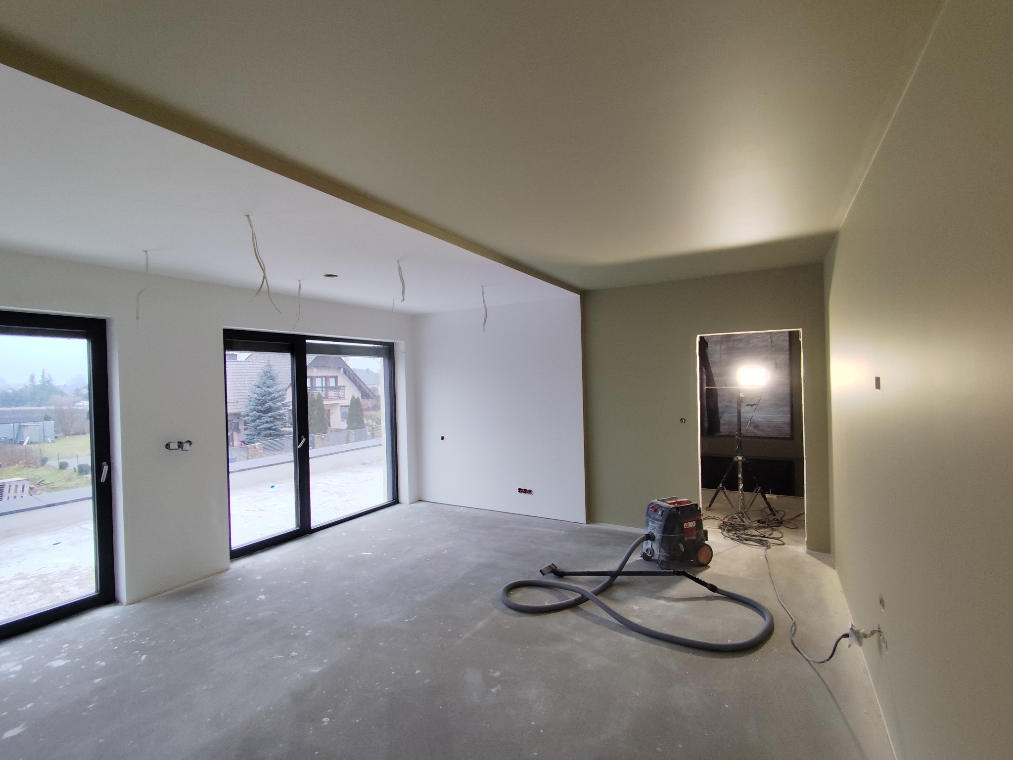 Wnętrze po szpachlowaniu: gładkie, białe ściany, duże okna z widokiem na zewnątrz, szpachlarka na podłodze. Przygotowanie do malowania.