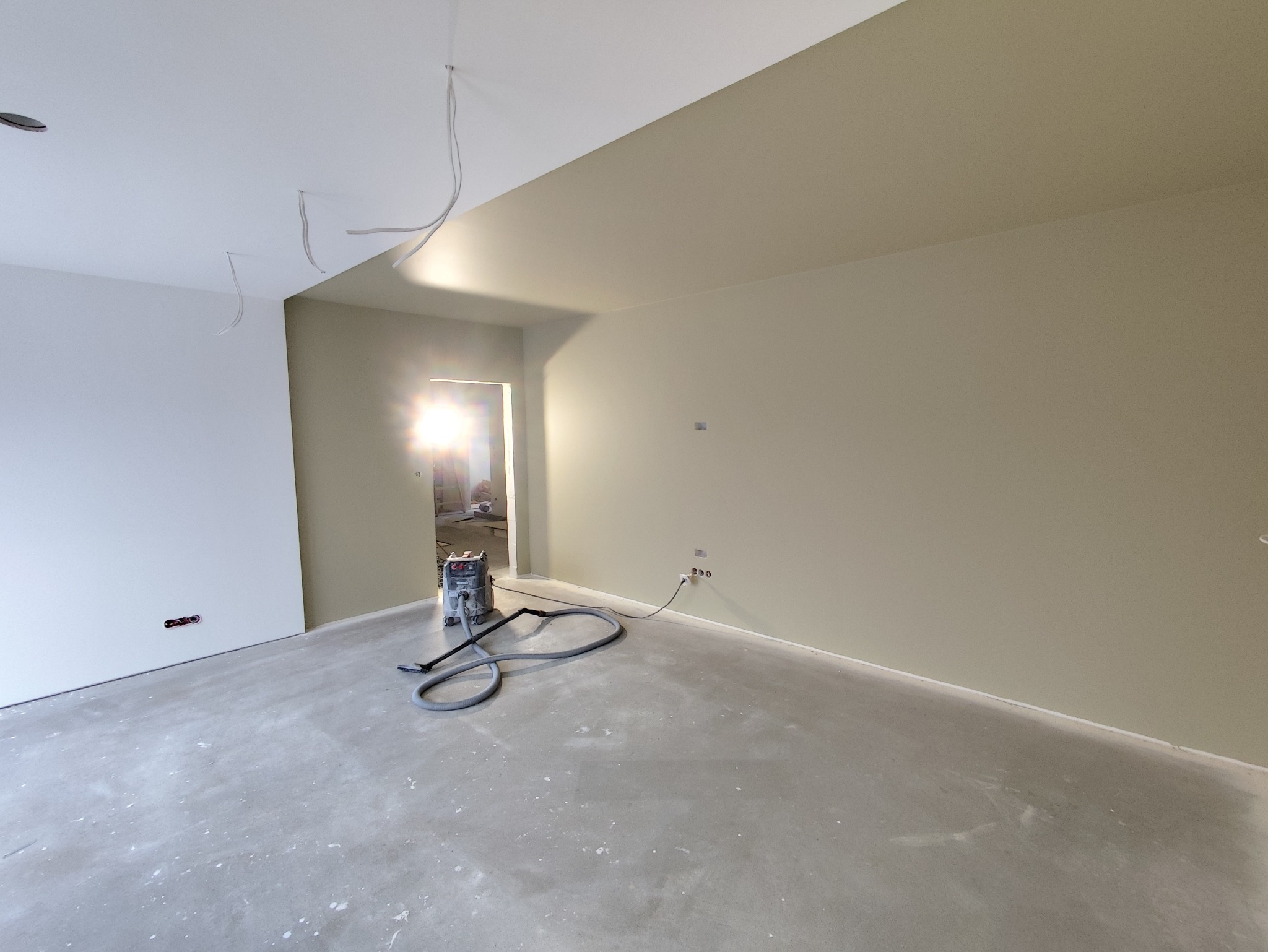 Wnętrze w trakcie prac wykończeniowych: ściany pomalowane na beżowo, widoczny agregat szpachlarski i kable zwisające z sufitu. Podłoga betonowa, przygotowana do dalszych prac.