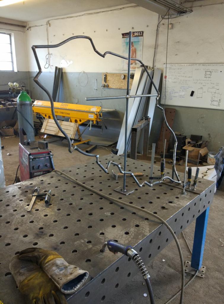 Metalowa konstrukcja przypominająca zarys postaci na perforowanym stole spawalniczym, z widocznym sprzętem spawalniczym, butlą z gazem i narzędziami w tle warsztatu.