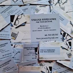 Stos wizytówek z napisem 'Usługi Księgowe mgr Anna Jacaszek', kalkulator i okulary na jasnym tle.