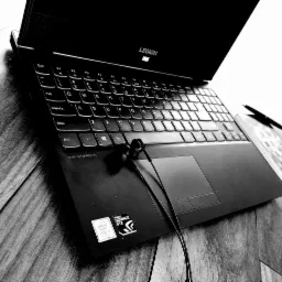 Czarno-białe ujęcie otwartego laptopa Lenovo Legion z słuchawkami na touchpadzie, długopisem i notatnikiem obok, na drewnianym biurku.