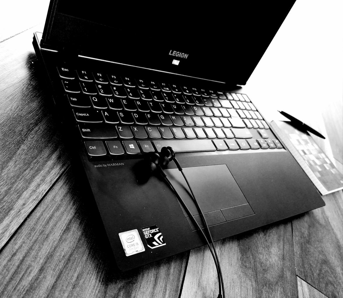 Czarno-białe ujęcie otwartego laptopa Lenovo Legion z słuchawkami na touchpadzie, długopisem i notatnikiem obok, na drewnianym biurku.