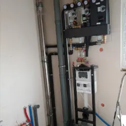 Wnętrze w trakcie montażu instalacji hydraulicznej i grzewczej, widoczne rury miedziane i plastikowe, rozdzielacze, manometry, zawory oraz stelaż podtynkowy WC KOLO, ściana z widocznymi śladami po...