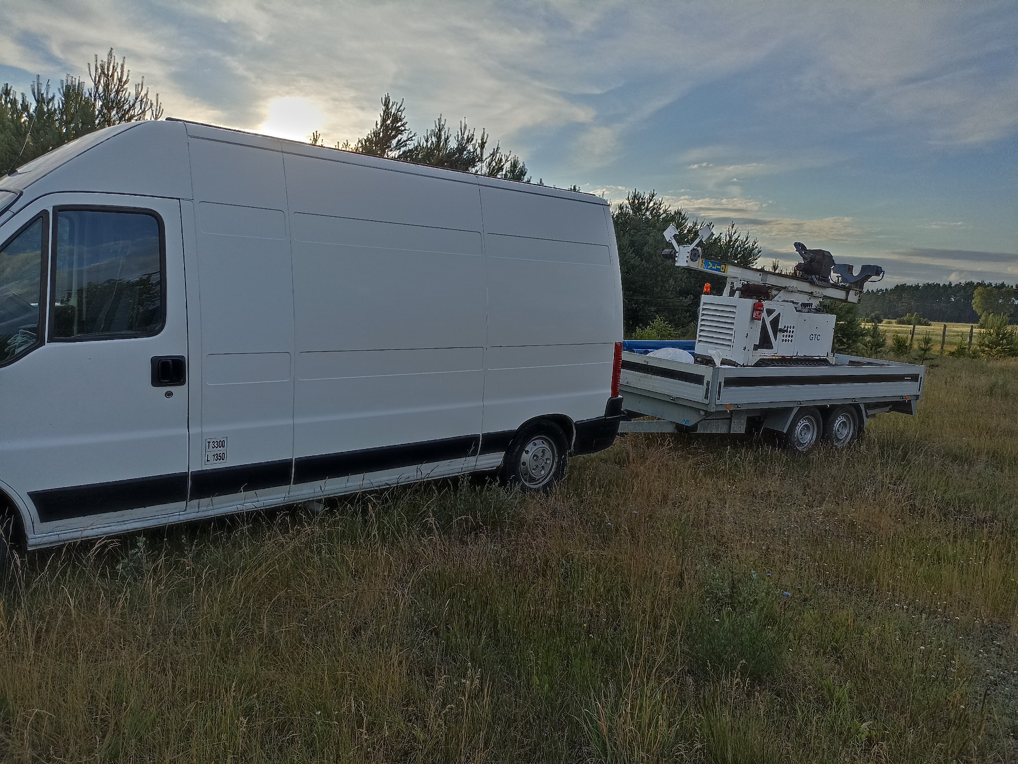 Biały bus z przyczepą, na której znajduje się urządzenie wiertnicze GTC, stoją na zarośniętym terenie, w tle widoczne drzewa i niebo z promieniami słonecznymi.