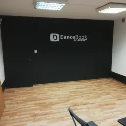 Pomalowana na czarno ściana z logo DanceBook Academy w pustym pomieszczeniu z panelami podłogowymi i krzesłami.