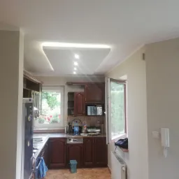 Kuchnia z ciemnymi drewnianymi szafkami, oświetlonym podwieszanym sufitem i oknem z widokiem na ogród, z niebieskim ręcznikiem na lodówce.