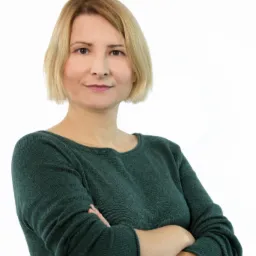 Portret kobiety z blond włosami do ramion, ubranej w ciemnozielony sweter, stojącej na białym tle z założonymi rękami.