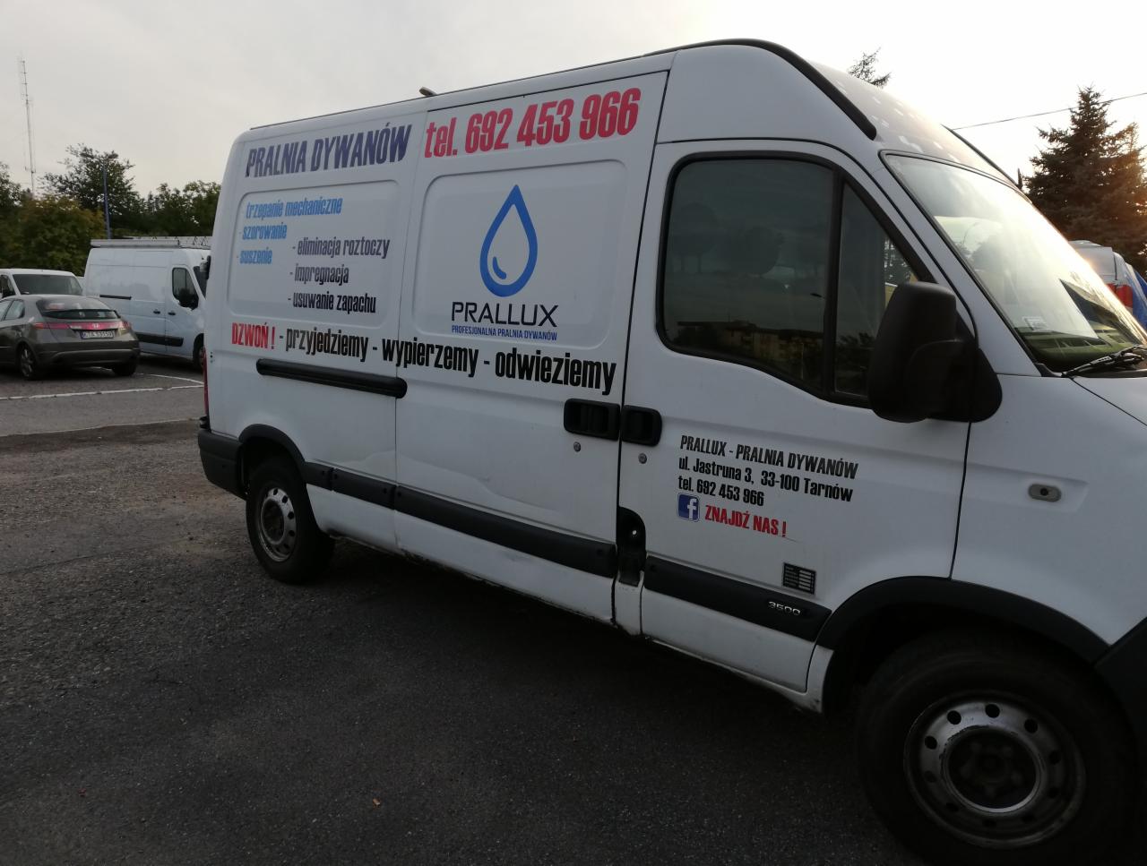 Biały samochód dostawczy z reklamą pralni dywanów Prallux, z numerem telefonu i hasłem 'Dzwon! Przyjedziemy - wypierzemy - odwieziemy', zaparkowany na parkingu.