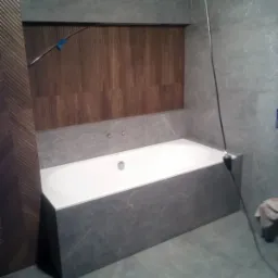 Nowoczesna łazienka z wanną obudowaną szarą płytką imitującą kamień, drewniana wnęka nad wanną, szare płytki na ścianach i podłodze, widoczne elementy instalacji.