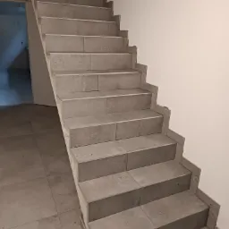 Schody wyłożone szarymi płytkami ceramicznymi, z widoczną podłogą również wyłożoną tymi samymi płytkami, biała ściana po prawej stronie i otwarte drzwi w tle