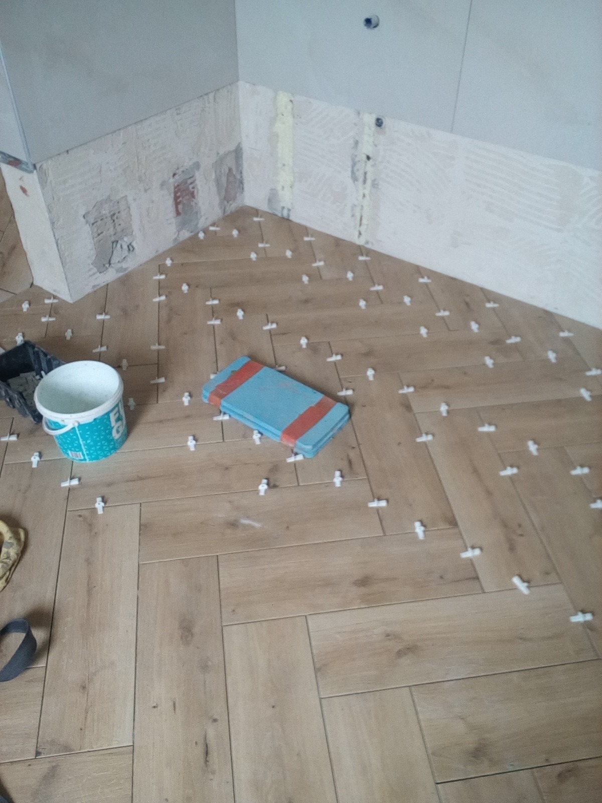 Układanie podłogi z płytek imitujących drewno w jodełkę, z użyciem krzyżyków dystansowych, widoczny fragment ściany przygotowanej do dalszych prac glazurniczych, pojemnik z klejem i gąbka.