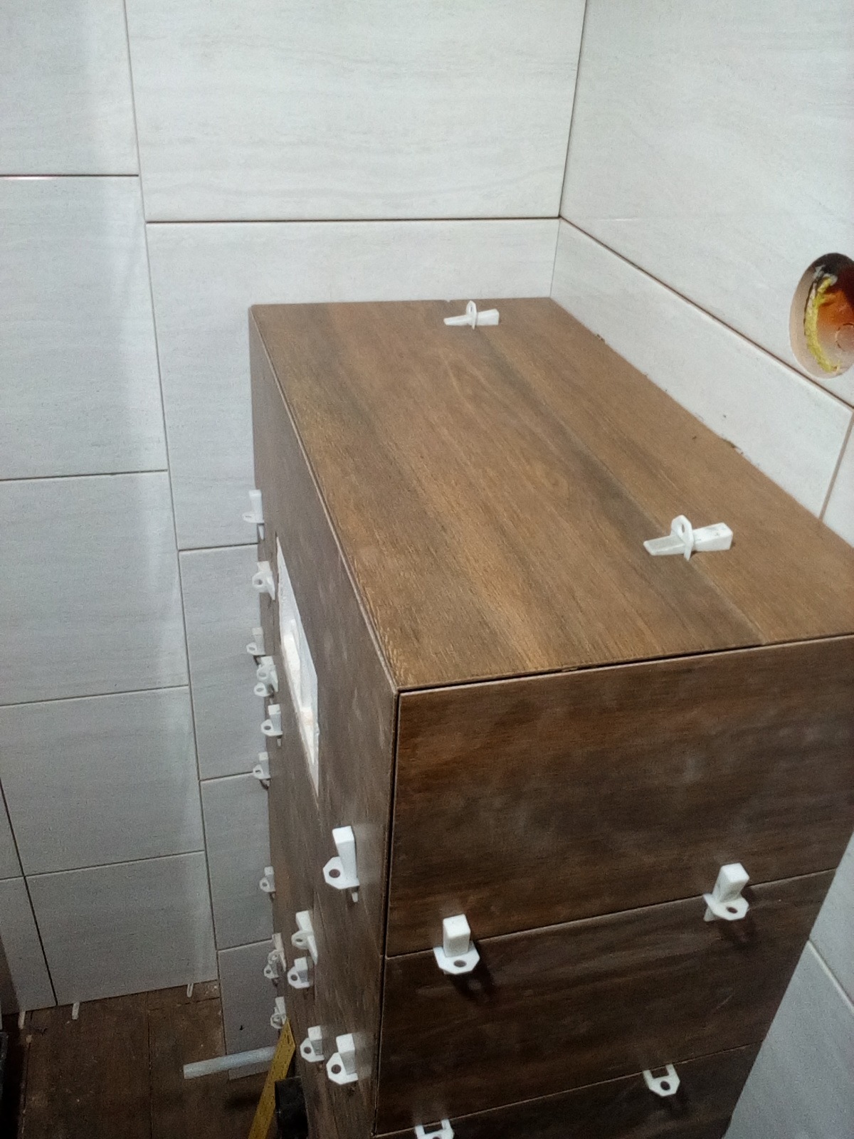 Obudowa WC w trakcie układania płytek imitujących drewno, z użyciem białych klipsów poziomujących; w tle jasne płytki ścienne.