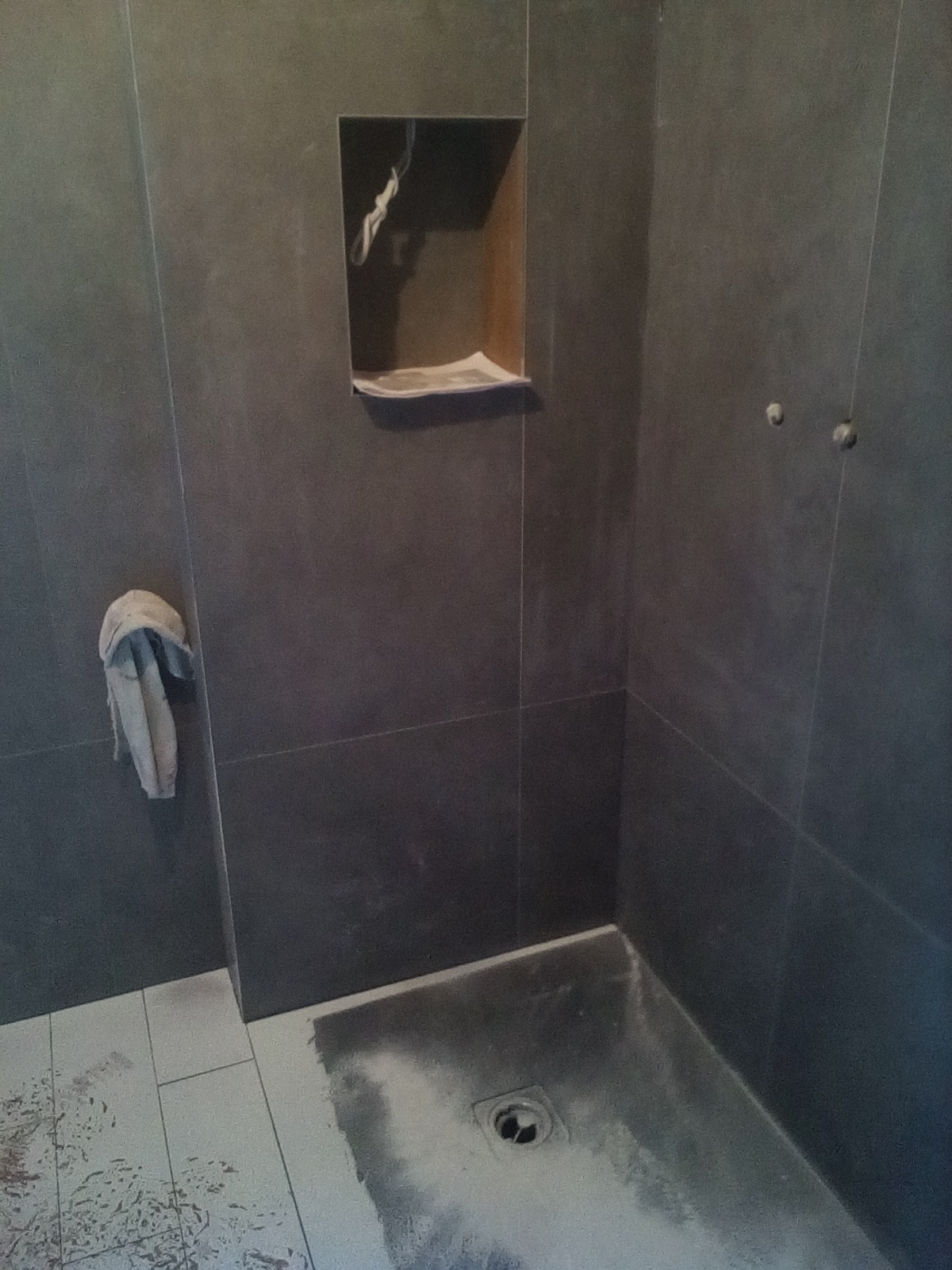 Niedokończona kabina prysznicowa z ciemnoszarymi płytkami, widoczna wnęka w ścianie, ręcznik na wieszaku i resztki zaprawy na podłodze.