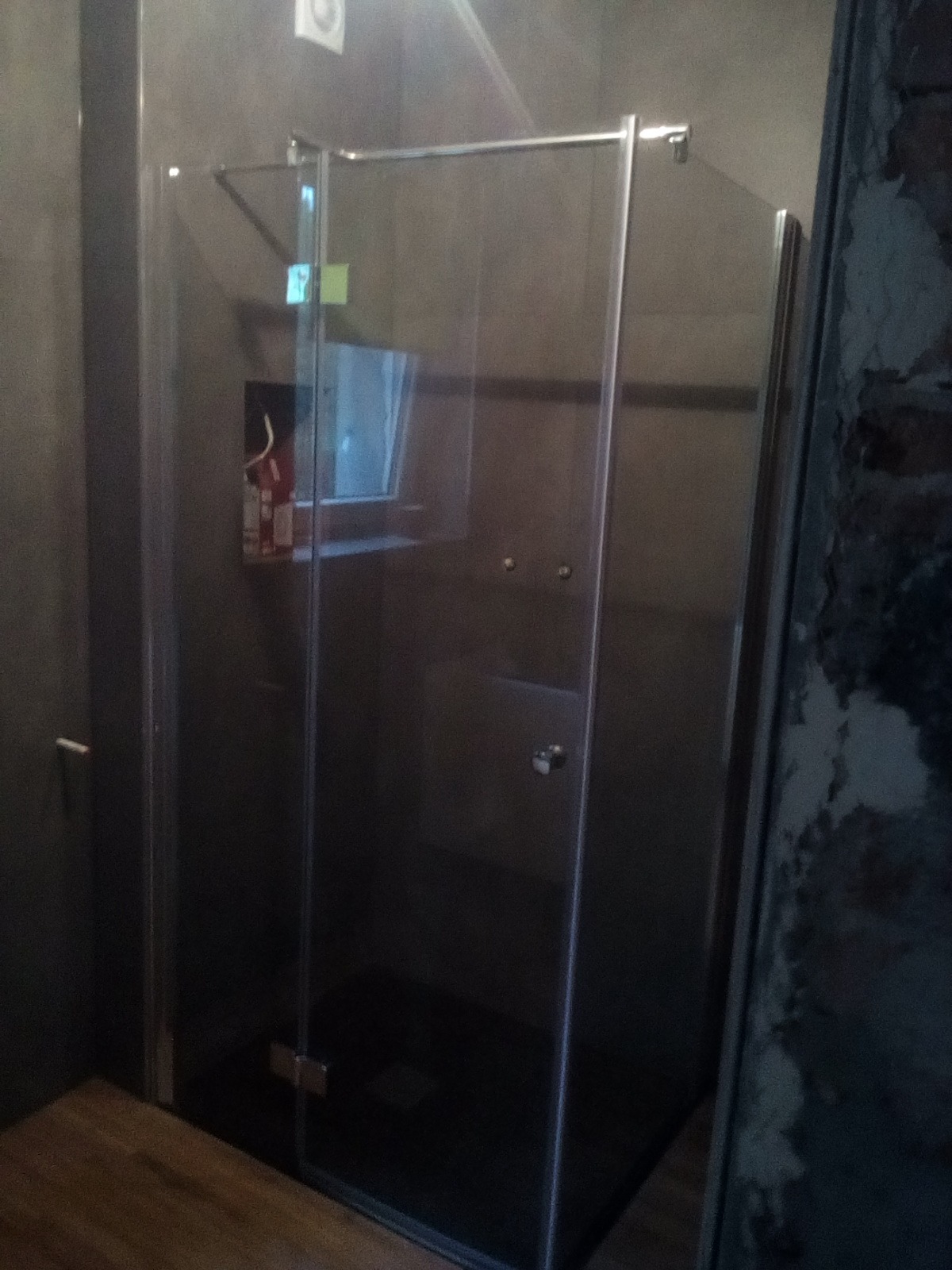 Szklana kabina prysznicowa z czarnym brodzikiem w łazience z szarymi ścianami, widoczny fragment drewnianej podłogi i fragment ściany z surową fakturą.