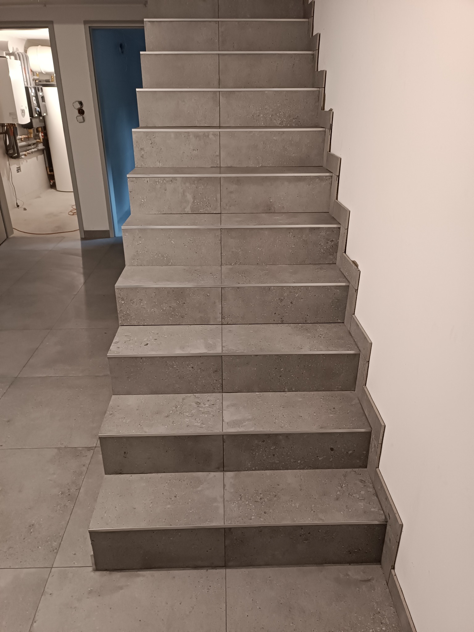 Schody wyłożone szarymi płytkami imitującymi beton, z metalowymi listwami na krawędziach stopni, widoczne z dołu, z otwartymi drzwiami do pomieszczenia technicznego w tle.
