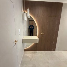 ADAL Sp. z o.o. - Nowoczesny korytarz: okrągłe, podświetlane lustro, minimalistyczna konsola, dwie lampy wiszące z mosiężnymi detalami. Ściana z drewnianymi panelami, ukryte drzwi. Jasne płytki.