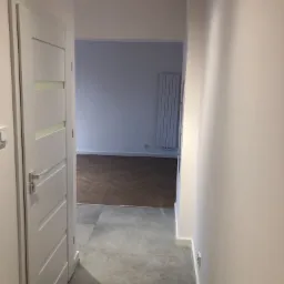 Otwarta biała, wewnętrzna stolarka drzwiowa z przeszkleniami, szara podłoga z gresu w korytarzu przechodząca w parkiet ułożony w jodełkę w pokoju z grzejnikiem panelowym na ścianie w tle.