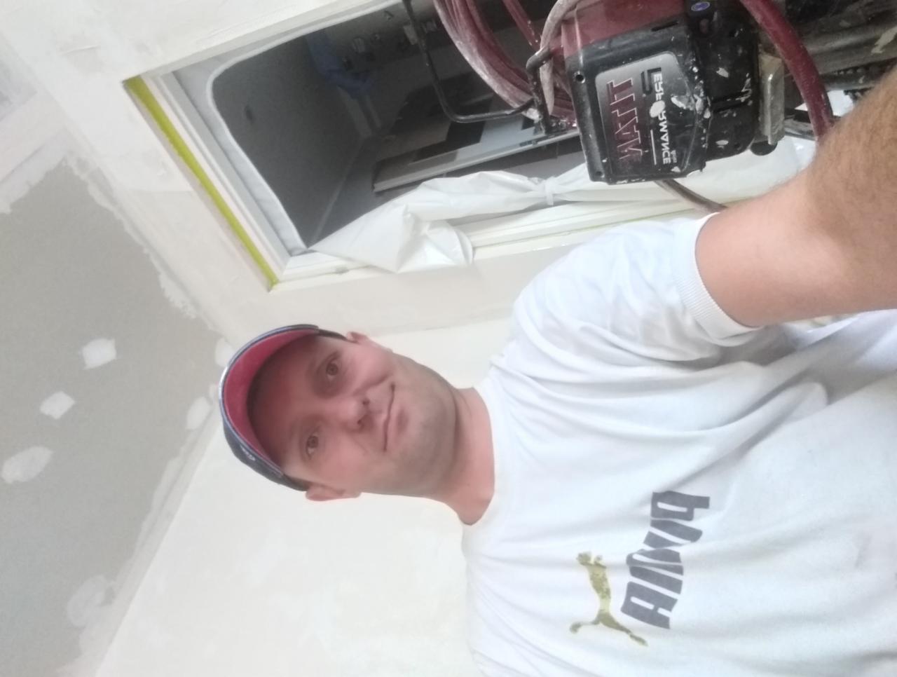 Uśmiechnięty mężczyzna w trakcie malowania wnętrza natryskowo. Widoczny fragment sufitu po szpachlowaniu i oklejone okno.