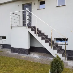 Zewnętrzne schody z ciemnymi płytkami i balustradą ze stali nierdzewnej, prowadzące do drzwi wejściowych domu z szarą elewacją.