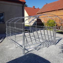 Formag - Gotowa, ocynkowana konstrukcja stalowa z rurek i siatki, prawdopodobnie paśnik dla zwierząt, ustawiona na żwirowym podłożu przed budynkami gospodarczymi z czerwonej cegły.