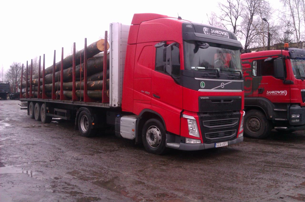 Czerwony samochód ciężarowy Volvo z załadowanym drewnem na platformie, zaparkowany na mokrym placu, z widocznym logo firmy transportowej Jankowski na kabinie.