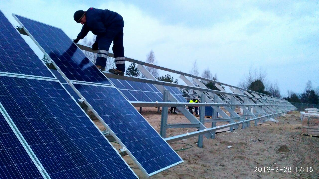 Montaż paneli słonecznych na metalowej konstrukcji; pracownik w kombinezonie i czapce instaluje panel na tle pochmurnego nieba i zimowego krajobrazu.