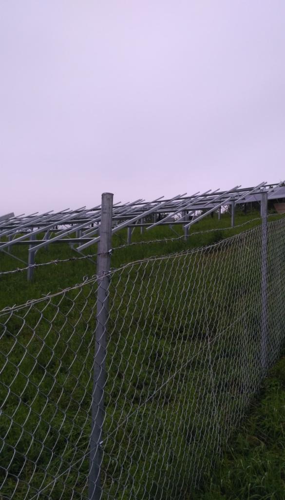 Stalowa konstrukcja wsporcza pod panele słoneczne na pagórkowatym terenie, widoczna przez siatkę ogrodzeniową z drutem kolczastym, pochmurne niebo w tle.
