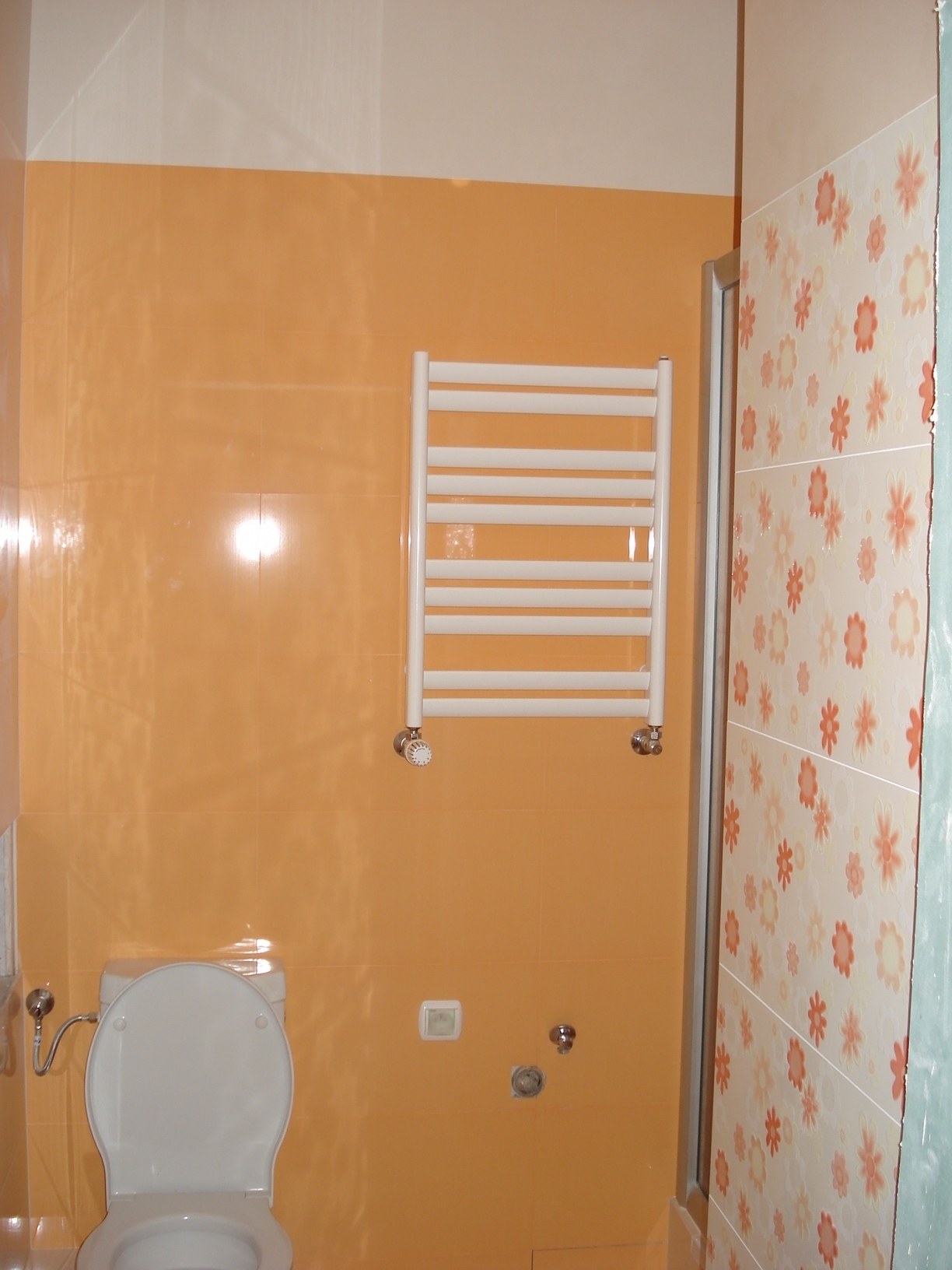 Pomarańczowa łazienka z grzejnikiem drabinkowym, toaletą i kwiatowymi płytkami w kabinie prysznicowej.
