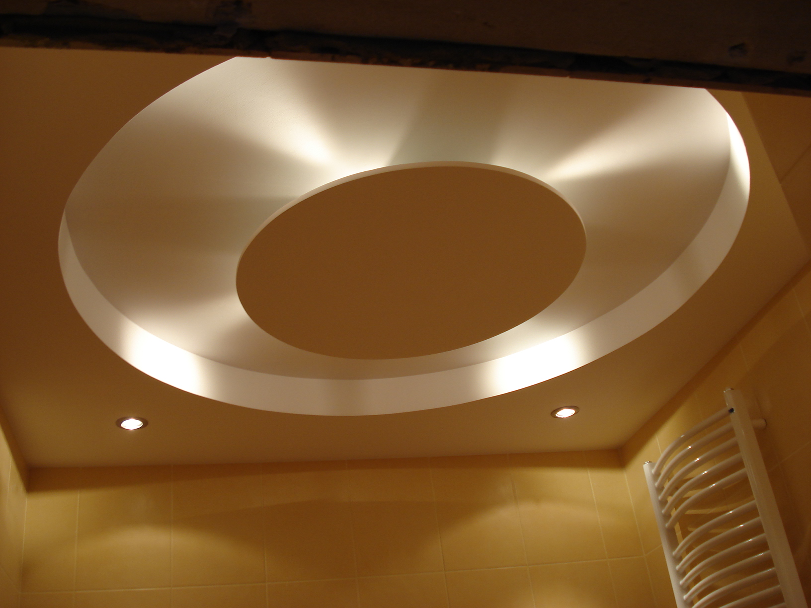 Okrągła, podwieszana konstrukcja sufitowa z oświetleniem punktowym w łazience wykończonej płytkami w odcieniach beżu.