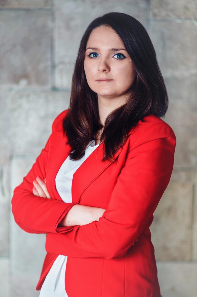 Portret kobiety w czerwonej marynarce z założonymi rękami na tle szarej ściany.