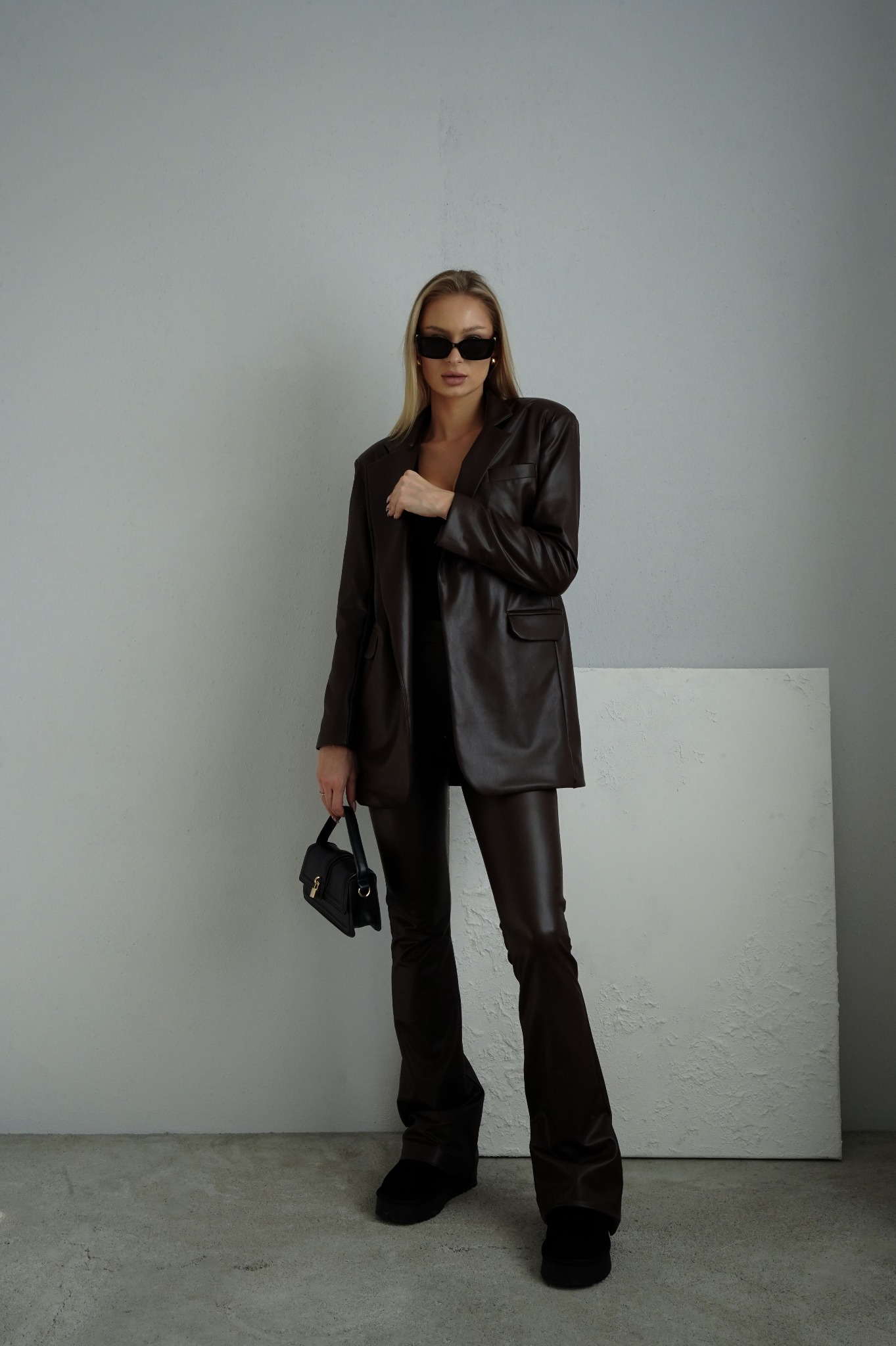 Modelka w brązowym skórzanym garniturze: oversize marynarka i spodnie dzwony, czarne okulary i torebka na tle szarej ściany i białego płótna.