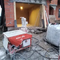 Czerwona maszyna do mieszania zaprawy 'mixer plus up' na budowie domu z cegły, widoczne przewody, worek z materiałem budowlanym i częściowo otynkowane wnętrze garażu z surową żarówką.