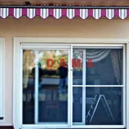 Zewnętrzna markiza w biało-bordowe pasy nad oknem z wewnętrznymi przesłonami i przesuwnymi drzwiami z siatką moskitierą oraz drabina w tle.