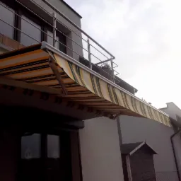 Zewnętrzna markiza w biało-żółto-zielone pasy zamontowana nad oknem i balkonem budynku mieszkalnego, widok z dołu.