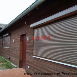 Brązowe rolety zewnętrzne zamontowane na drewnianym domu w Chojnicach, widoczne trzy okna z roletami i drzwi wejściowe.