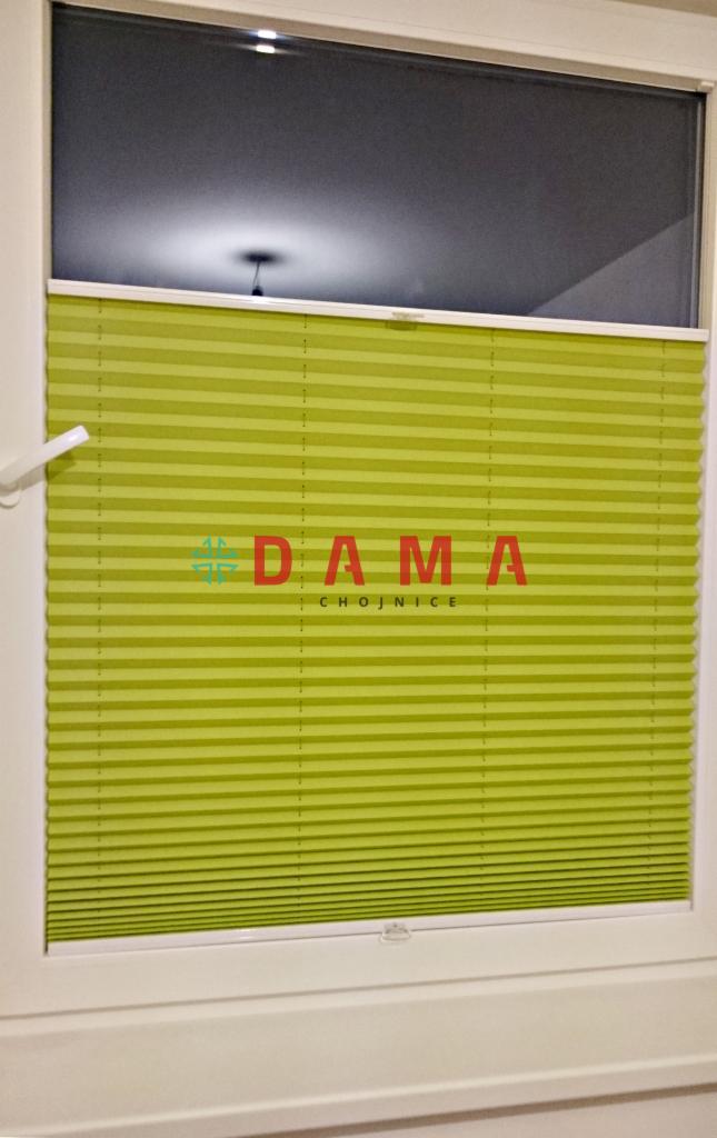 Zamknięta, jasnozielona osłona okienna typu harmonijka z widocznym logo 'DAMA Chojnice' na tle szyby okiennej.
