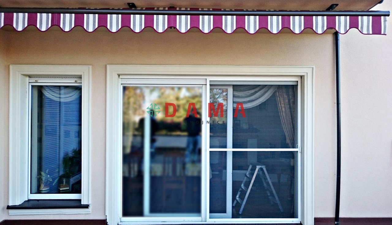 Zewnętrzna markiza w biało-bordowe pasy nad oknem z wewnętrznymi przesłonami i przesuwnymi drzwiami z siatką moskitierą oraz drabina w tle.