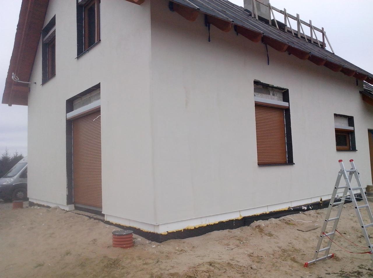 Nowy dom jednorodzinny z zamontowanymi brązowymi osłonami okiennymi, widoczne rusztowanie na dachu i drabina oparta o ścianę, teren wokół domu w trakcie prac wykończeniowych.
