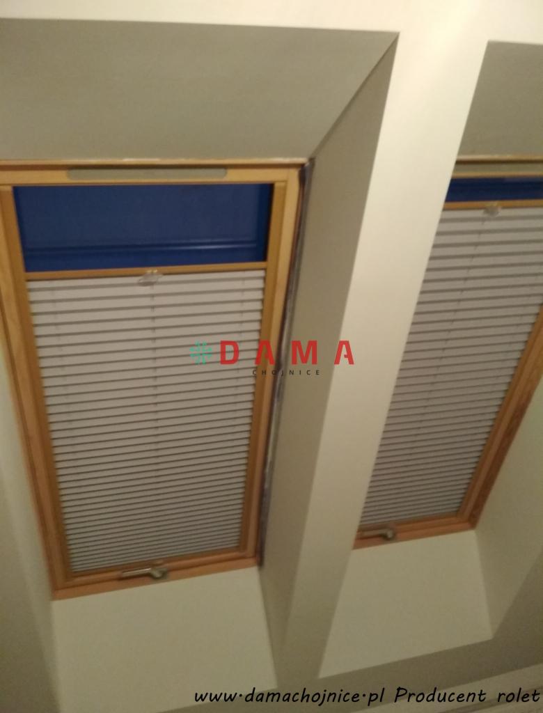 Dwa okna dachowe z drewnianą ramą, wyposażone w szare plisy częściowo zasłonięte, z widocznym niebieskim fragmentem górnej części okna, w tle biała skośna ściana poddasza.