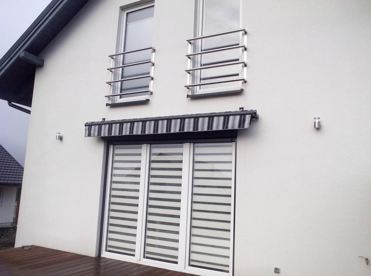 Elewacja domu z oknami zabezpieczonymi metalowymi balustradami, nad tarasowym oknem markiza w czarno-białe pasy, a w oknie na parterze zamontowane poziome przesłony okienne.