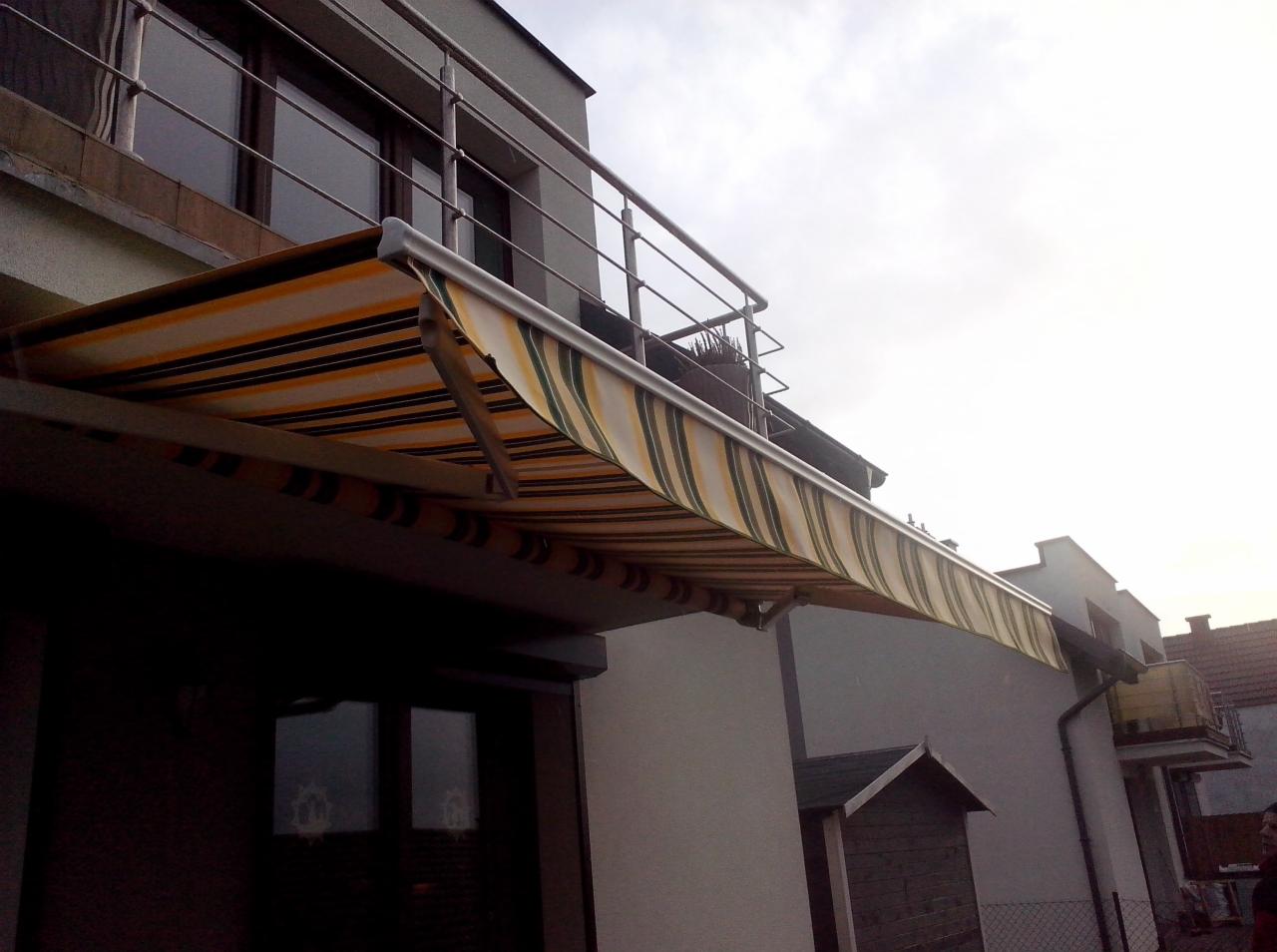 Zewnętrzna markiza w biało-żółto-zielone pasy zamontowana nad oknem i balkonem budynku mieszkalnego, widok z dołu.