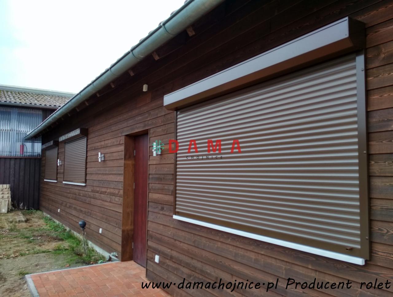 Brązowe rolety zewnętrzne zamontowane na drewnianym domu w Chojnicach, widoczne trzy okna z roletami i drzwi wejściowe.