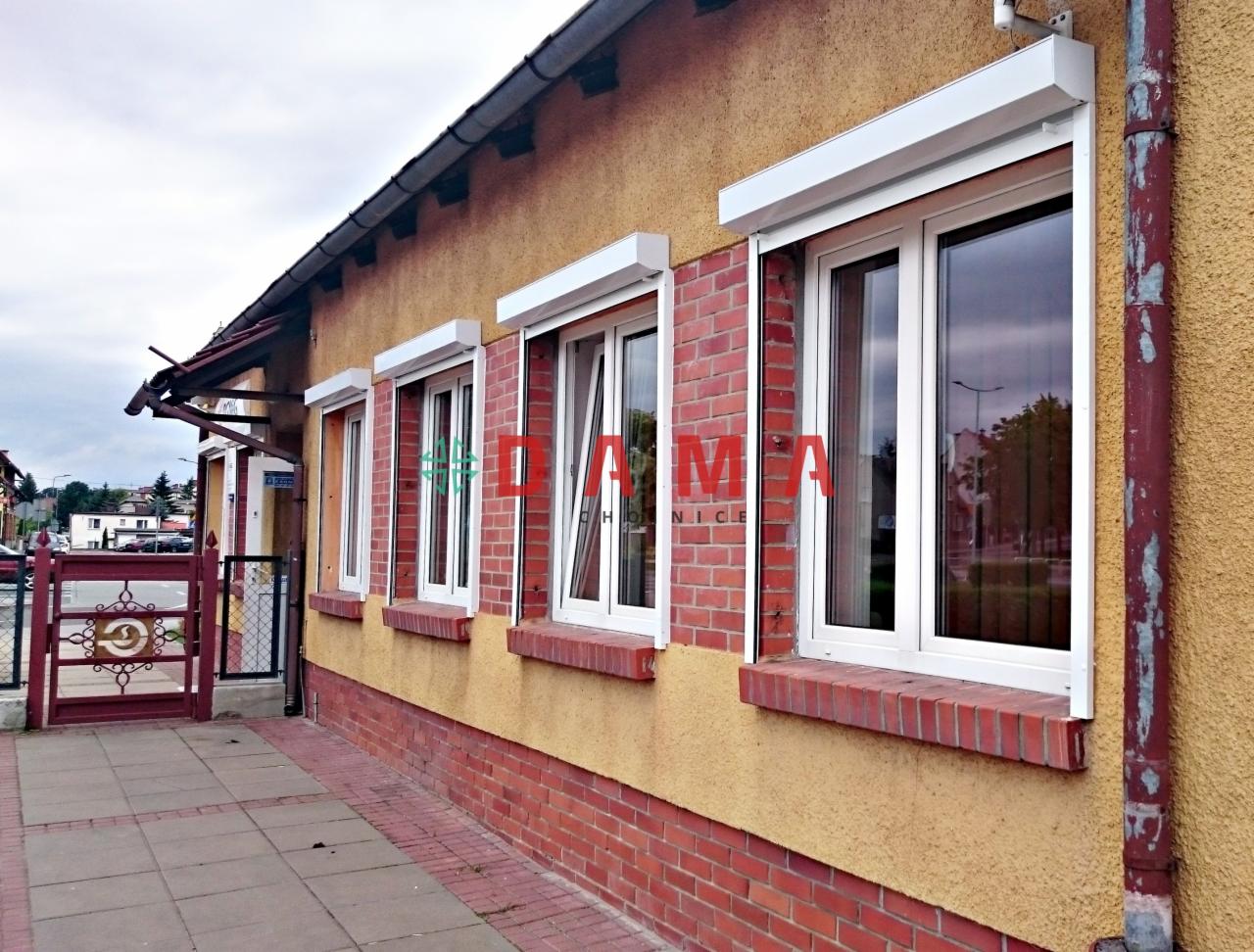 Żółta elewacja budynku z czerwonymi ceglanymi detalami i białymi oknami, na których zamontowano białe kasety osłonowe.