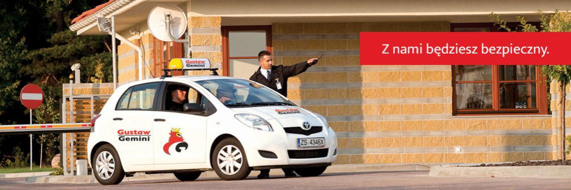 Samochód firmy ochroniarskiej Gustav Gemini z pracownikiem wskazującym kierunek, brama wjazdowa i budynek w tle, napis 'Z nami będziesz bezpieczny' w prawym górnym rogu.