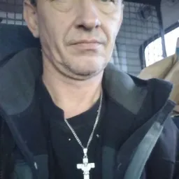 Portret mężczyzny w czarnej kurtce i z srebrnym łańcuszkiem z krzyżem na tle wnętrza samochodu dostawczego.