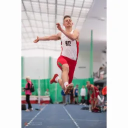 Skok w dal: atletyczny mężczyzna w locie, numer startowy 972, czerwono-biały strój, hala sportowa, dynamiczne ujęcie, widoczne detale butów i napięcie mięśni.