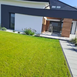 Nowoczesny dom jednorodzinny z białymi ścianami i grafitowymi elementami, widoczna drabina oparta o ścianę przy wejściu, zadbany trawnik i elementy dekoracyjne z kamienia.