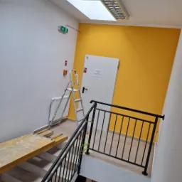 Klatka schodowa po odświeżeniu koloru ścian na żółty, widoczna drabina malarska i rusztowanie, białe ściany i sufit z oknami dachowymi.